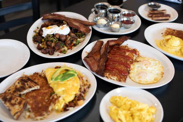 Large breakfast spread featuring avocado omelettes, crispy hash browns, thick-cut bacon, scrambled eggs, and roasted potato hash with poached eggs — a classic American diner feast with a variety of savory options and sides.