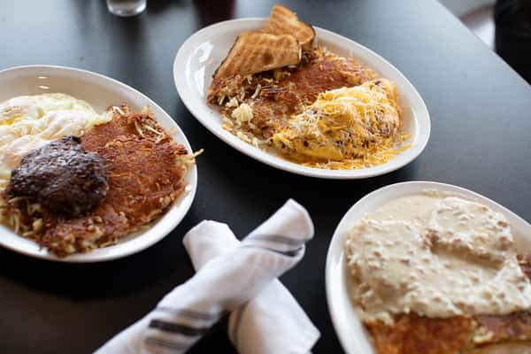 Hearty American breakfast plates with crispy hash browns, fluffy scrambled eggs topped with cheddar cheese, sausage patties, over-easy eggs, and biscuits smothered in creamy country gravy — served in a casual diner setting.