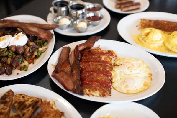 Crispy hash browns topped with ketchup, paired with sunny-side-up eggs and thick-cut bacon slices. Nearby dishes include eggs Benedict, breakfast sausage, and a vibrant vegetable hash, creating a full and indulgent diner-style breakfast spread.