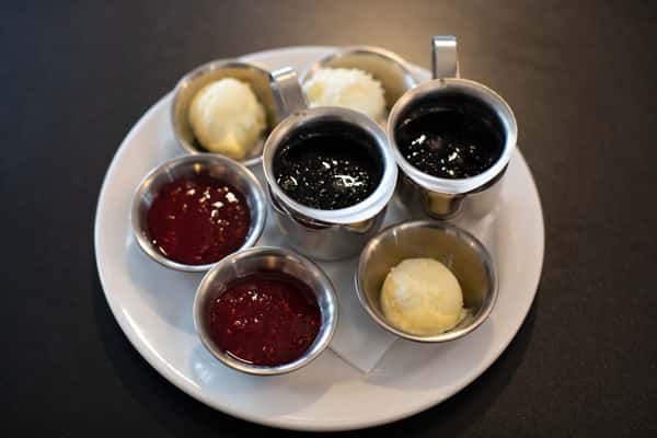 Assorted breakfast condiments on a white plate, featuring stainless steel cups filled with raspberry and blackberry jam, served alongside scoops of creamy butter — ideal for pancakes, waffles, or toast at a classic American diner.