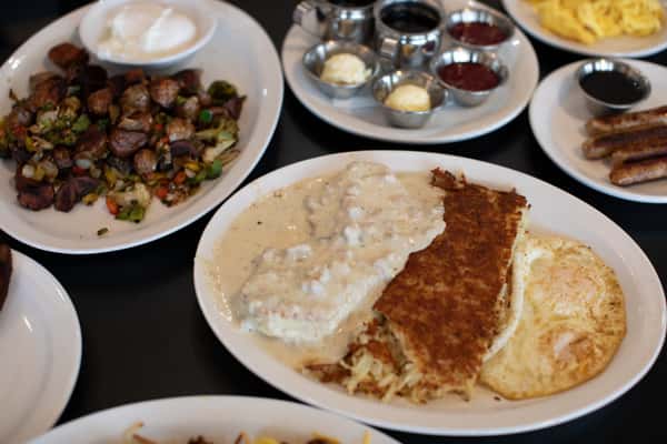 Classic Southern-style breakfast with biscuits smothered in creamy sausage gravy, served alongside golden hash browns and a sunny-side-up egg. Surrounding plates include roasted breakfast potatoes with sour cream and a variety of jams and butters for a hearty diner meal.