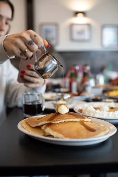 Golden buttermilk pancakes stacked high and topped with a scoop of butter, being generously drizzled with maple syrup — a comforting and timeless breakfast favorite in a bustling diner setting.