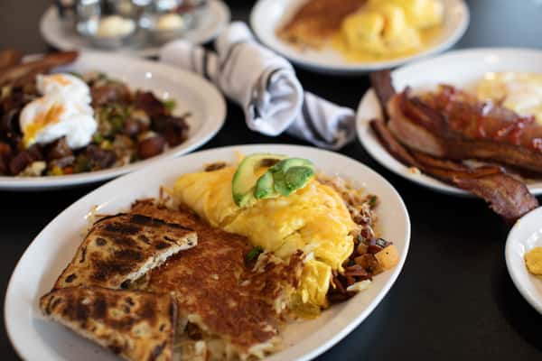 Classic American diner breakfast featuring a fluffy omelette topped with avocado slices, served with crispy hash browns, grilled toast, and hearty sides including bacon, eggs, and roasted breakfast potatoes — perfect for a satisfying morning meal.