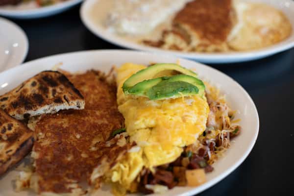 A breakfast plate with a fluffy omelet topped with sliced avocado, crispy hash browns, shredded cheese, and breakfast potatoes, with toasted flatbread on the side and other breakfast plates blurred in the background.
