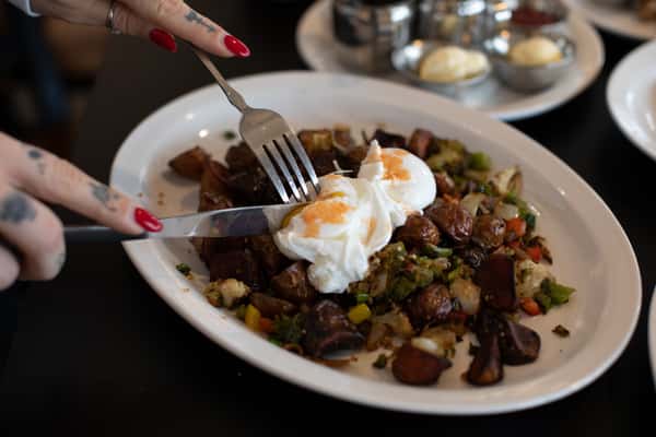 Close-up of a rustic breakfast hash being sliced into, topped with perfectly poached eggs and surrounded by roasted vegetables — a fresh, flavorful option at a homestyle breakfast diner.