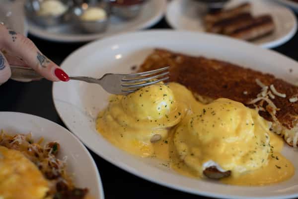 Close-up of eggs Benedict being sliced into, revealing rich yolks beneath creamy hollandaise sauce. Served with crispy hash browns at a traditional American breakfast diner.