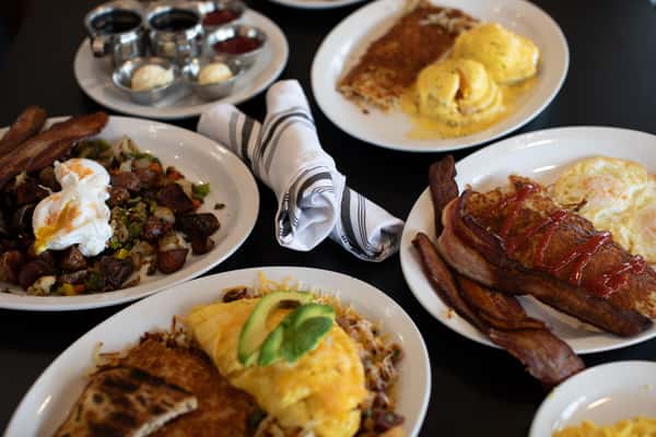 A table filled with breakfast dishes, including eggs Benedict, an omelet topped with avocado, crispy bacon, breakfast potatoes with poached eggs, and pancakes or French toast with syrup, arranged on white plates.