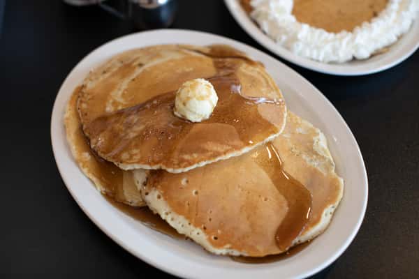 Golden stack of fluffy buttermilk pancakes topped with a swirl of butter and drizzled with maple syrup, with a playful whipped cream smiley pancake in the background — classic and fun diner breakfast.