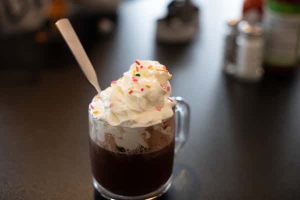 Rich hot chocolate topped with whipped cream and colorful rainbow sprinkles, served in a clear glass mug — a cozy and festive drink option at a family-friendly breakfast spot.