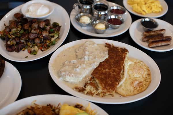 Generous American diner breakfast spread featuring biscuits smothered in sausage gravy, crispy hash browns, sunny-side-up eggs, sautéed breakfast potatoes with peppers, scrambled eggs, sausage links, and a side of assorted jams and butter — served on white plates for a hearty and classic morning meal.