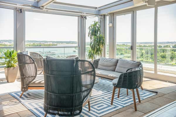 Indoor-outdoor restaurant seating area with dark wicker chairs, a gray sofa, and a patterned rug, with large windows overlooking the waterfront