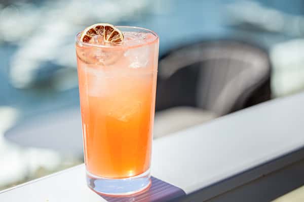 Orange cocktail garnished with a dried citrus wheel in a highball glass, on a table at a restaurant with a patio view in the background