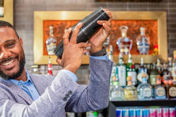 Bartender shaking a cocktail shaker behind the bar, with bottles of liquor on shelves in the background at a restaurant
