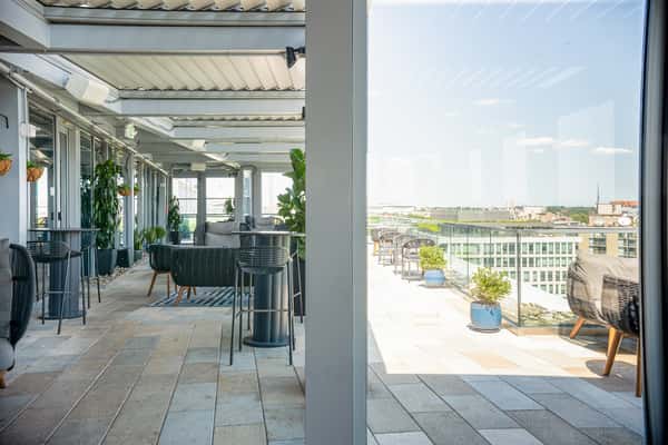 Rooftop patio dining area with outdoor seating, potted plants, and pergola structures, overlooking a waterfront cityscape at a restaurant