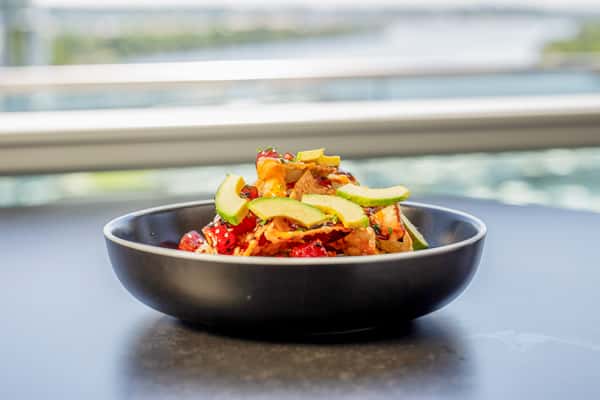 Tuna nachos with crispy chips, avocado, and tomatoes in a dark bowl on a table, with a window and outdoor view in the background