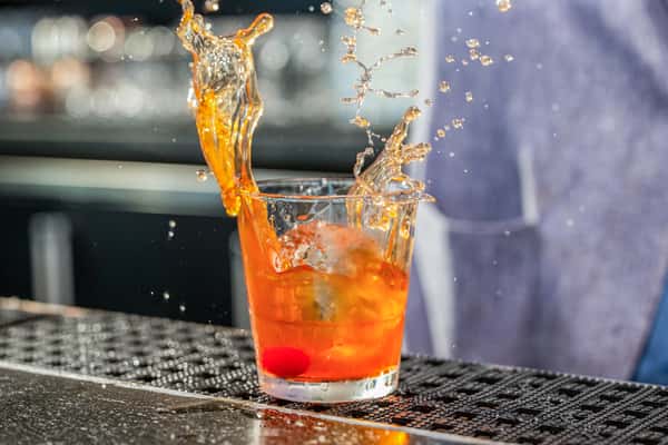 Orange cocktail with a dramatic splash effect and a cherry garnish in a rocks glass with ice, on a metal bar counter at a restaurant