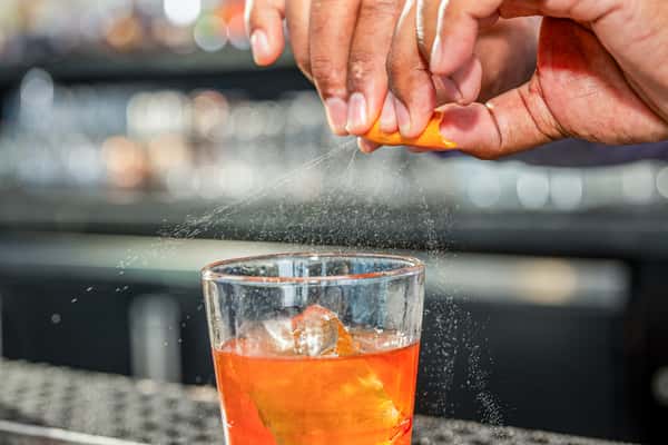 Hand squeezing a citrus peel over an orange cocktail with ice in a rocks glass, on a bar counter at a restaurant
