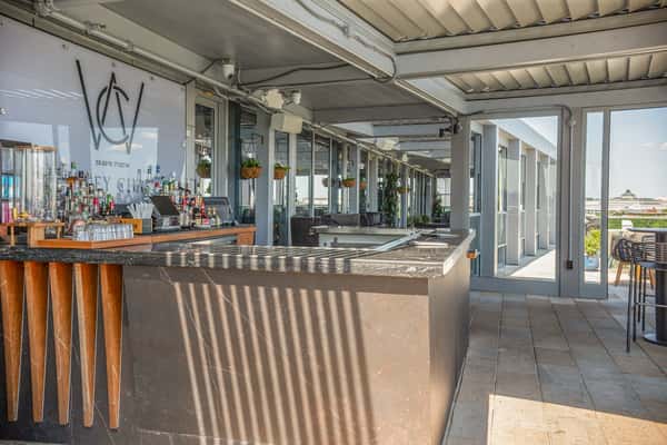 Outdoor bar area with a wooden counter, string lights, and bottles displayed on shelves, under a covered patio structure at a restaurant