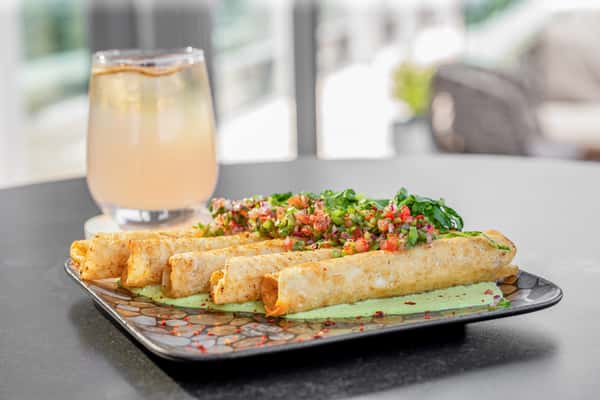 Crispy chicken taquitos on a rectangular plate at a restaurant, with a cocktail visible in the background