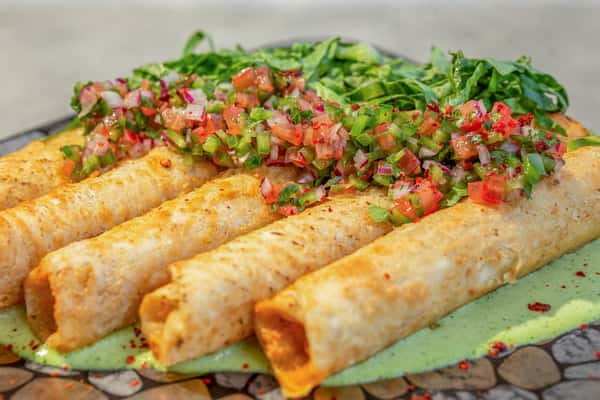 Crispy chicken taquitos topped with pico de gallo, served on a bed of shredded greens on a patterned plate at a restaurant