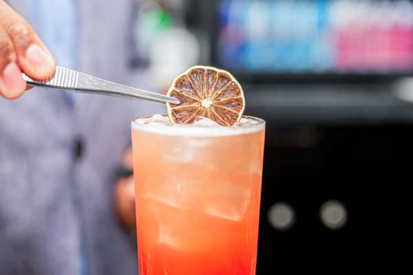 Hand holding an orange cocktail garnished with a dried citrus wheel in a highball glass, outdoors at a restaurant
