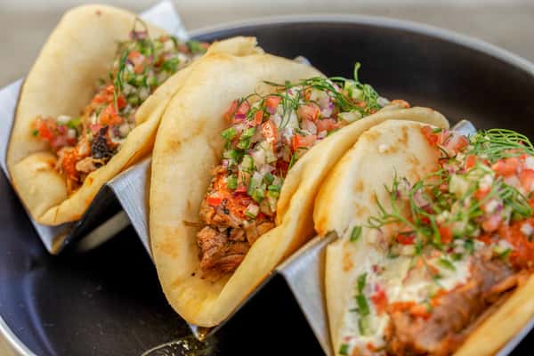Lamb shawarma tacos with meat, fresh herbs, and diced vegetables in soft shells, standing upright on a dark plate at a restaurant