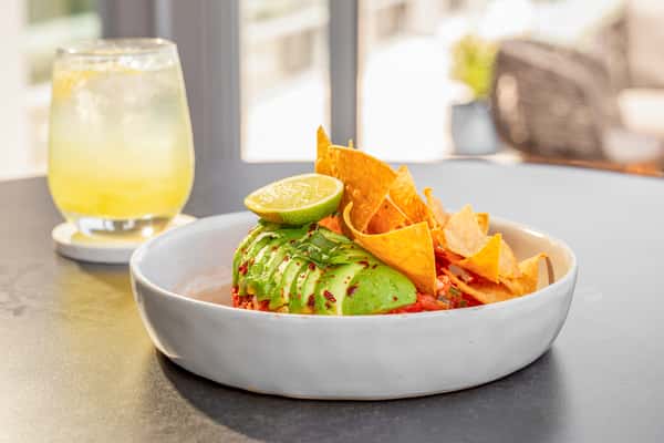 Shrimp ceviche with sliced avocado and tortilla chips on a white plate, with a cocktail visible in the background at a restaurant