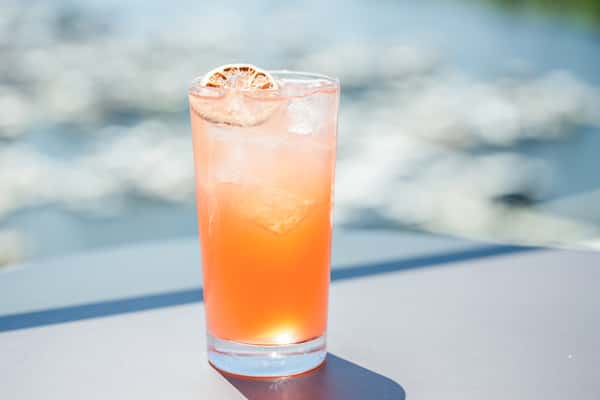 Orange gradient cocktail garnished with a dried citrus wheel in a highball glass, outdoors with a blurred waterfront view