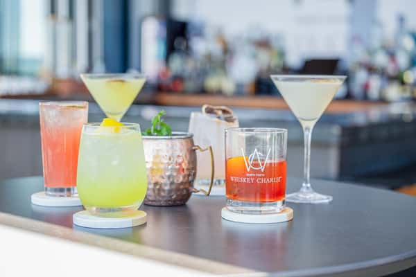 Lineup of assorted cocktails including martinis, a yellow citrus drink, a Moscow Mule in a copper mug, and a branded rocks glass on a bar at a restaurant