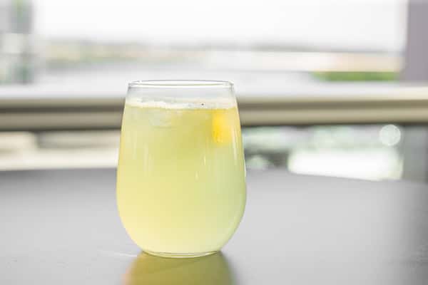 Yellow citrus drink with a lemon slice and ice in a stemless wine glass, with a waterfront view in the background at a restaurant