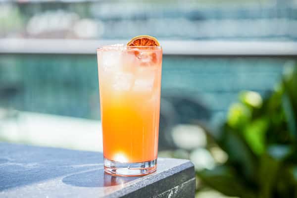Orange gradient cocktail garnished with a dried citrus wheel in a highball glass, resting on a stone ledge outdoors at a restaurant