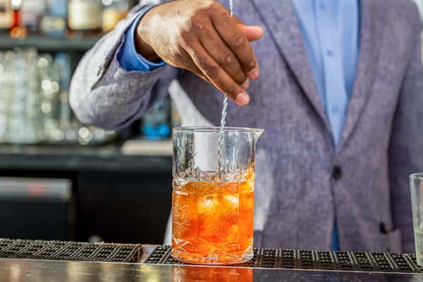 Bartender's hand garnishing an orange cocktail in a mixing glass with ice, behind the bar at a restaurant
