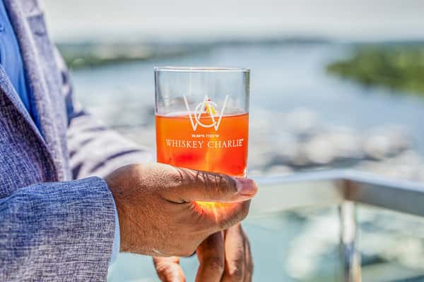 Hand holding a branded rocks glass with an orange cocktail, outdoors with water in the background at a restaurant