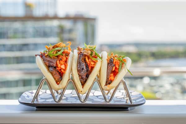 Brisket bao buns with pickled vegetables and scallions on a metal stand, with a waterfront and city buildings in the background