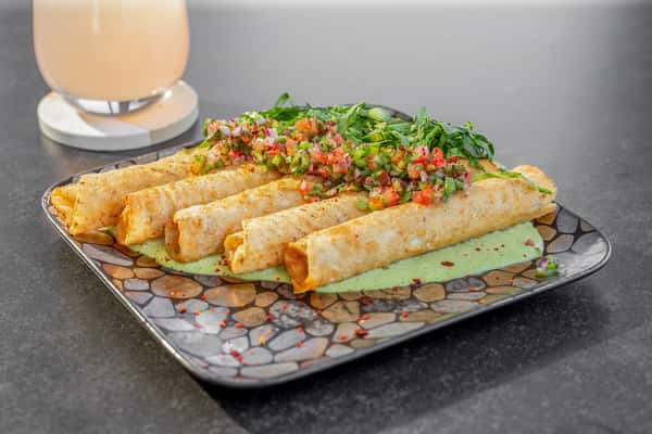 Crispy chicken taquitos topped with pico de gallo, served on a stone-patterned plate with a candle in the background at a restaurant
