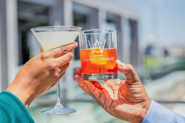 Two hands toasting with a martini glass and a rocks glass containing an orange cocktail, outdoors at a restaurant