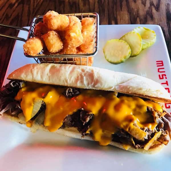 Cheesesteak sandwich topped with American cheese and a side of pickles and tater tots