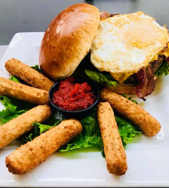 Burger with an egg, baconm and cheese on w brioche bun with a side of mozzerella sticks on a bed a lettuce
