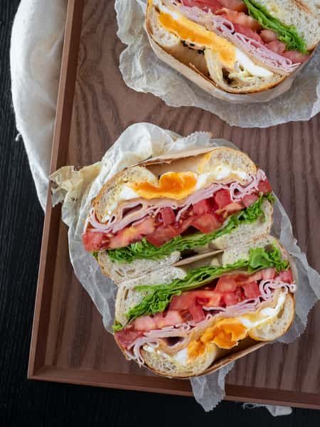 Ham, egg, and tomato sandwich on artisan bread with lettuce and soft-boiled egg yolk, served on parchment paper.