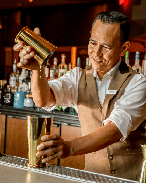 Bartender pouring a cocktail between golden shakers behind a well-stocked bar.