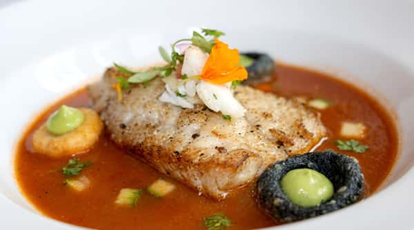 Pan-seared fish fillet in red broth, garnished with herbs, daikon, and an orange flower.