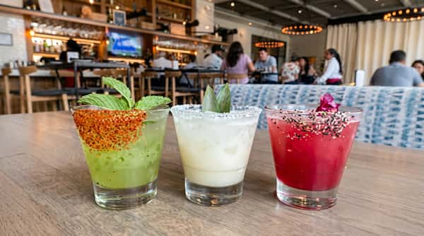 Three margaritas: green with chili-lime rim, white with salt rim, and red with a floral garnish.