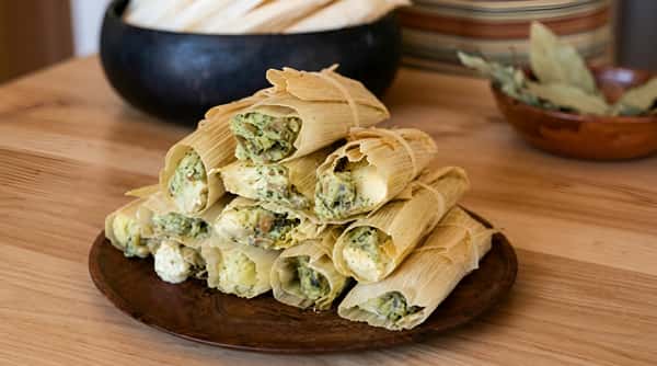 A pile of green chile and cheese tamales wrapped in corn husks on a brown plate.