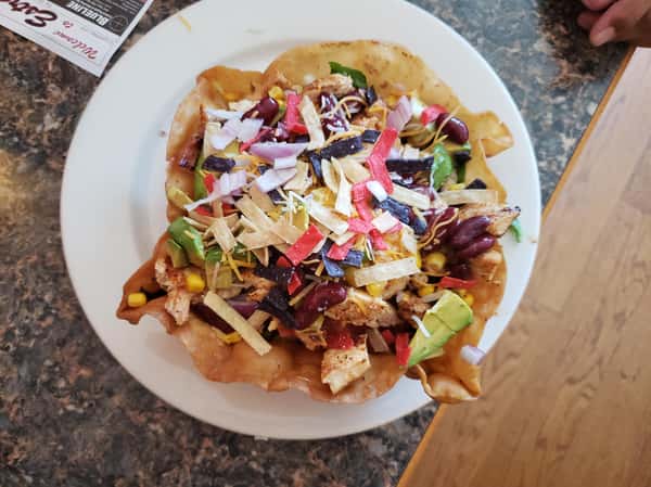 top view of the Chicken Avocado Taco Salad which includes Lettuce, tomato, cucumbers, corn, beans, cilantro & pepper jack cheese, Served in a hard shell tortilla with sour cream & guacamole
