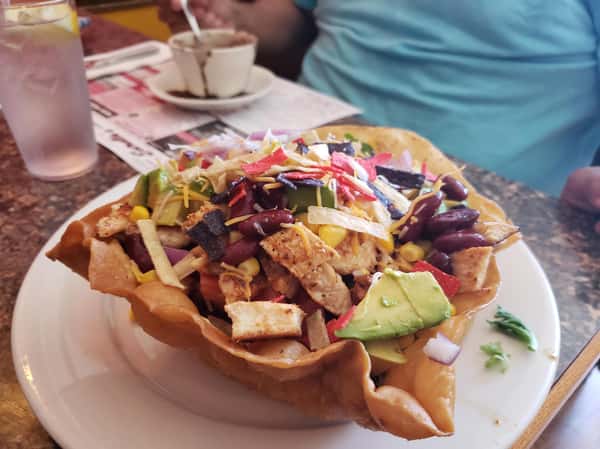 side view of the Chicken Avocado Taco Salad which includes Lettuce, tomato, cucumbers, corn, beans, cilantro & pepper jack cheese, Served in a hard shell tortilla with sour cream & guacamole