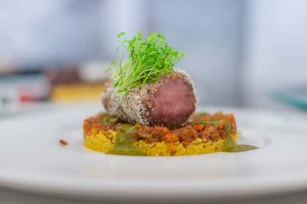 Sliced medium-rare beef topped with fresh microgreens, served over a colorful vegetable base with a golden sauce on a white plate.