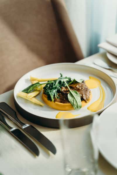 Elegantly plated gourmet dish featuring fresh green vegetables, herbs, and a golden sauce in a white ceramic bowl, served on a restaurant table.