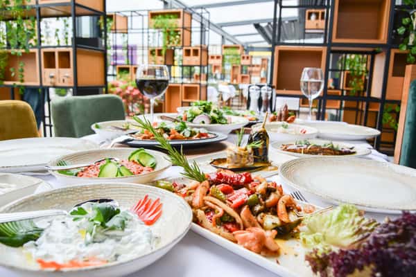 A beautifully presented selection of fresh dishes, including colorful salads and roasted entrees, arranged on a restaurant table with a bright, modern dining area in the background.