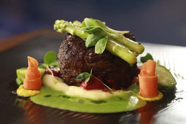Gourmet seared beef entrée topped with fresh green vegetables, served with a vibrant green puree, roasted vegetables, and colorful garnishes on a dark plate.
