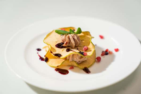 Elegantly presented gourmet dish featuring tender slices of meat layered over a golden base, garnished with fresh herbs, colorful vegetables, and decorative sauce accents on a white plate.